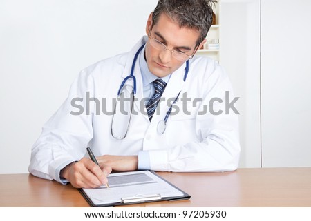 Male Doctor writing on clipboard sitting at desk.
