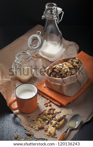 Top view on the packaged peanut cookies, broken biscuits, cups and jars of milk on paper and a dark wooden background.