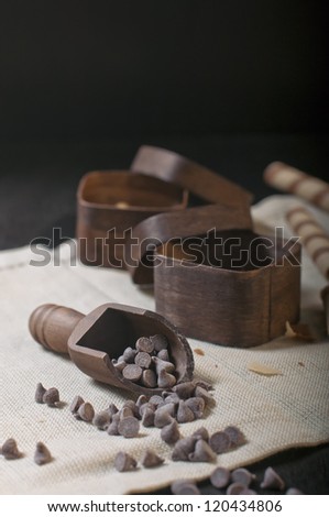 Side closeup view of wooden scoop with chocolate drops and two boxes on beige napkin and dark background.
