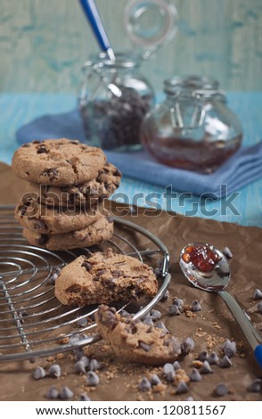 Side view of cookies with chocolate drops on round iron stand and brown paper, jars with jam and chocolate on cracked blue background