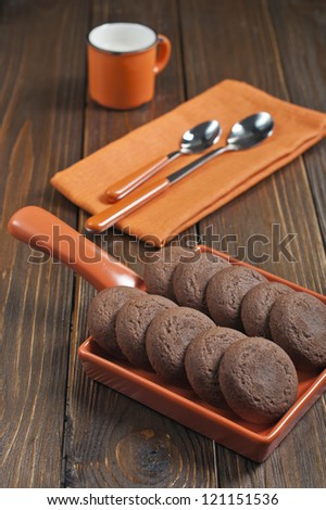 Biscuits in orange ceramic pan, mug of milk, spoons on napkin on dark wooden background