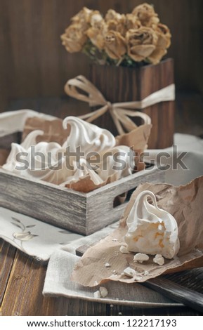 Side view of half of meringue and crumbs on paper, wooden box with meringues and dry roses in square vase on dark wooden background