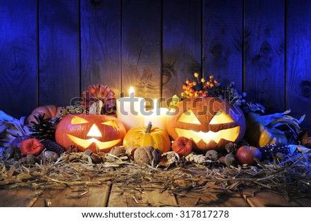 illuminated halloween pumpkins, candles, nuts, maize-cob and apple on straw in front of old weathered wooden board in blue light