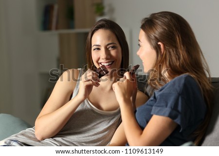 Two friends enjoying eating chocolate in the night sitting on a couch in the living room at home