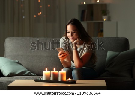Relaxed girl using phone in the night with candle lights sitting on a couch in the living room at home