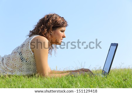 Profile of a beautiful woman lying on the grass browsing a laptop with a blue sky in the background