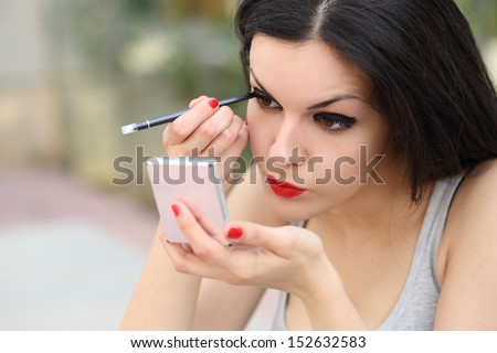 Beautiful woman making up eyelashes herself with a hand mirror outdoor
