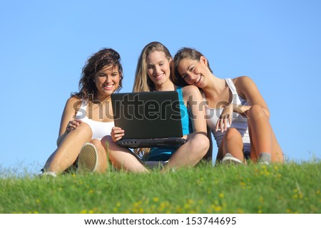Group Of Three Teenager Girls Laughing While Watching The Laptop Sitting On The Grass With The Sky In The Background