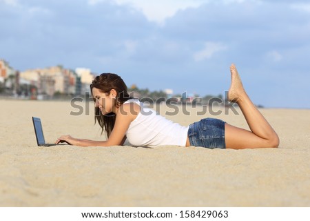 Profile of a teenager girl browsing her laptop lying on the sand of the beach with a city in the background