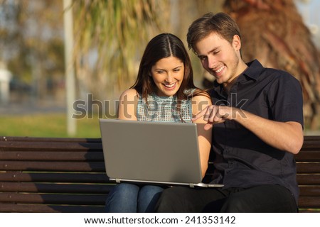 Happy couple laughing and watching media in a laptop sitting on a bench in a park