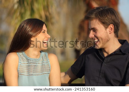 Friends or couple laughing and taking a conversation sitting on a bench in a park