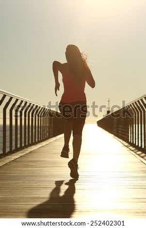 Back view of a runner silhouette running fast at sunset on a bridge on the beach
