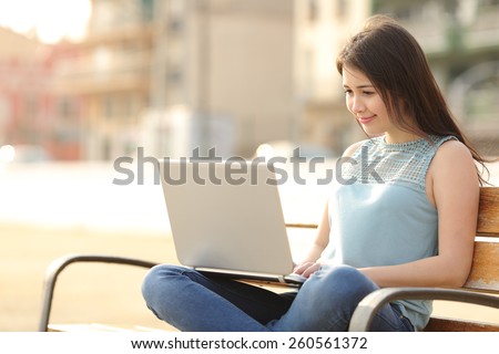 Student girl browsing a laptop and studying sitting in a bench in a park