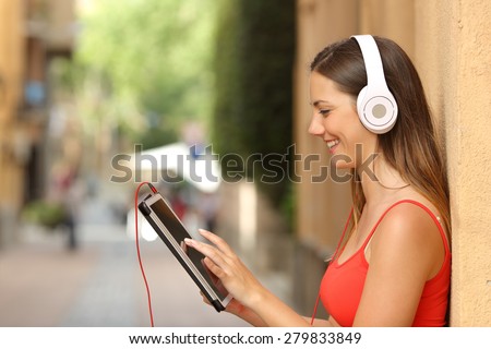 Happy girl browsing a tablet and listening to the music with headphones leaning on a wall in the street