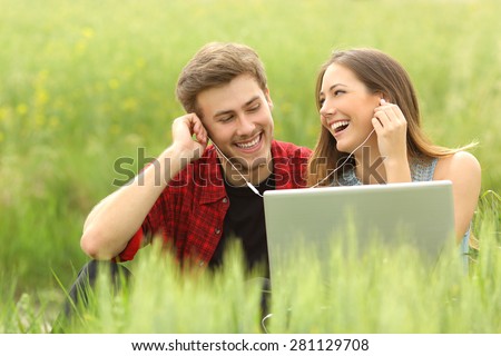 Happy couple or friends sharing music from a laptop sitting in a green field and looking each other