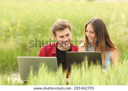 Couple or friends watching apps in two laptops in a green field