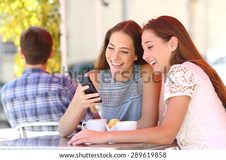 Two happy friends or sisters sharing a smart phone in a coffee shop terrace looking at device in summer