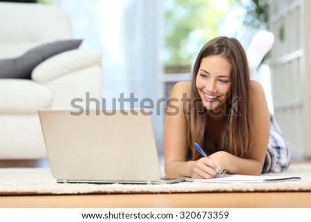 Student learning online with a laptop and taking notes lying on a carpet in the living room at home