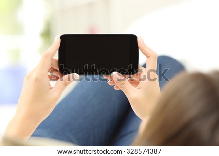 Back view of a close up of a woman hands watching media in a smart phone lying on a couch at home