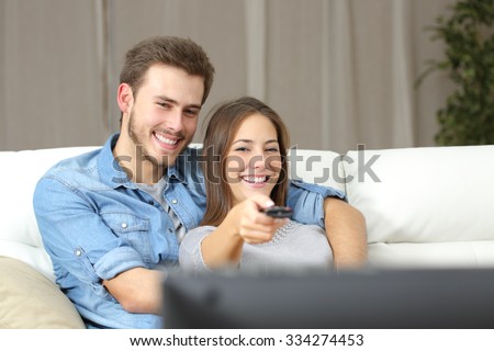 Happy couple using remote control to change channel on tv sitting on a couch at home