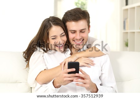 Funny couple or marriage sharing a smart phone to watch media content sitting on a couch at home