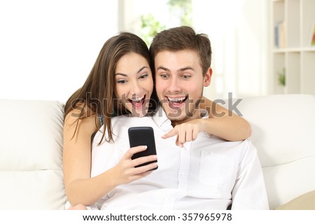 Attractive surprised and amazed couple watching smart phone together sitting on a couch in a living room at home