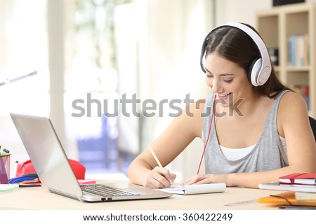 Portrait of a student learning on line with headphones and laptop taking notes in a notebook sitting at her desk at home