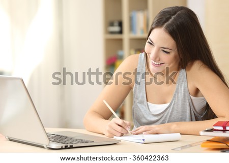 Happy student learning on line and taking notes in a notepad doing homework looking at laptop screen in a desk at home