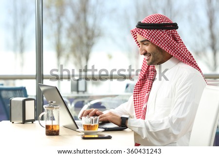 Arab saudi man working online with a laptop and smartwatch in a coffee shop or an hotel bar with a window and outdoor terrace in the background