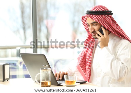 Arab man working on the phone and a laptop in a coffee shop with a window in the background