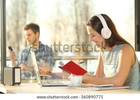 Profile of female listening music with headphones working on line writing notes in agenda in a coffee shop with people outside in the background