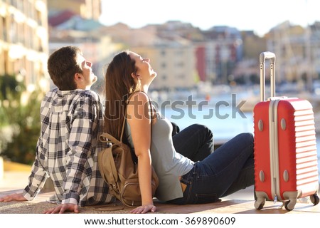 Side view of a couple of 2 tourists with a suitcase sitting relaxing and enjoying vacations in a colorful promenade. Tourism concept