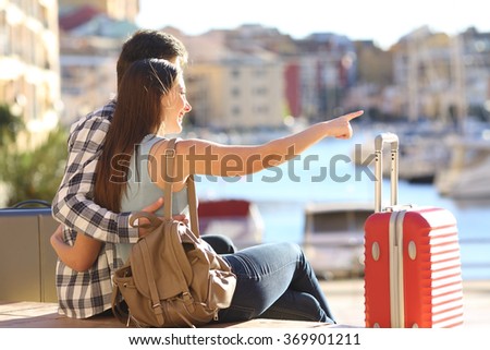 Portrait of couple of tourists with suitcases sitting and cuddling pointing at horizon on vacations in a port of a beach urbanization