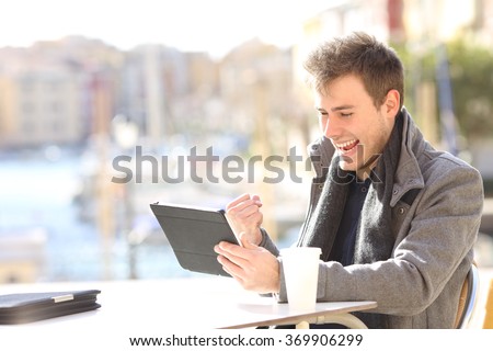 Handsome euphoric winner winning and watching a tablet in a coffee shop terrace of a port of urbanization with the sea in the background