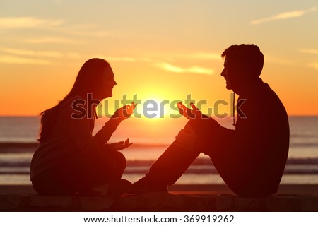 Side view of a full body of two friends or couple silhouette of teens sitting and talking at sunrise on the beach with the sun in the middle