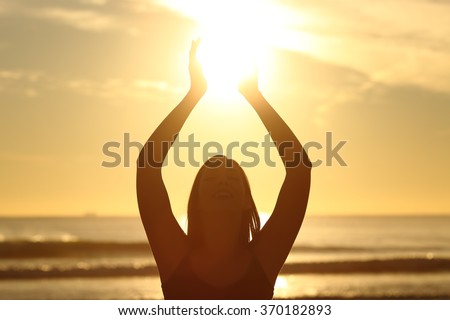 Front view of a back light of faithful woman silhouette holding sun on the beach at sunrise with a warm background