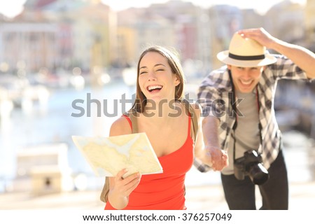 Front view of a happy couple of tourists running towards camera holding a paper map in a travel destination port with the sea in the background. Tourism concept