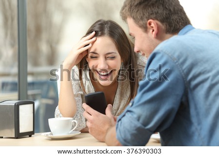 Cheerful couple laughing watching media in smart phone sitting in a coffee shop