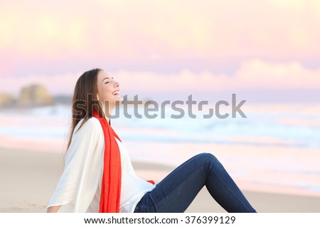 Side view of a happy woman sitting on the sand of the beach breathing fresh air at sunset with the ocean and a warmth pink sky in the background