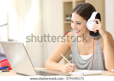Happy student studying watching video lecture on line and listening with headphones and taking notes sitting in a desktop at home
