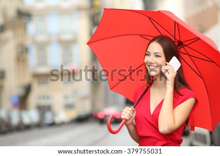 Happy woman in red talking on a smart phone under an umbrella in the street of an old town