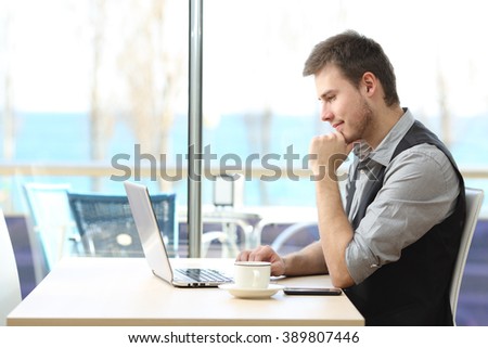 Side view of a businessman using a laptop on line sitting in a bar with a window and the sea in the background