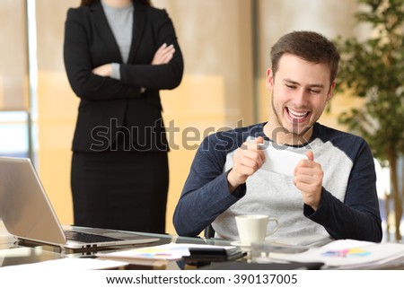 Lazy employee playing games with his smartphone sitting in a desktop while his angry boss is watching at office