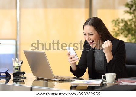Excited businesswoman winning after achievement reading a smart phone sitting in a desktop at office
