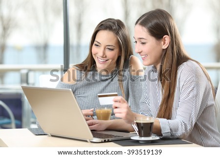Two friends buying on line with a laptop and a credit card in a coffee shop or hotel with the sea outdoors in the background