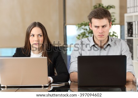 Front view of two angry businesspeople using computers disputing at workplace and looking sideways each other with envy