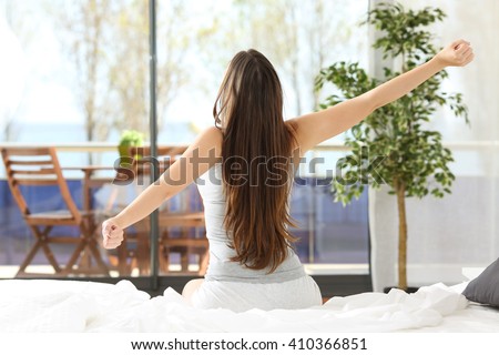 Woman stretching arms and waking up sitting on the bed in an hotel or home bedroom looking the sea outdoors through the window