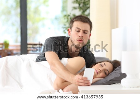 Jealous husband spying the phone of his partner while she is sleeping in a bed at home