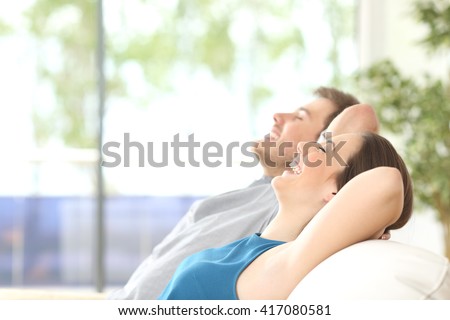 Side view of a happy couple breathing and resting lying in a couch at home with a window in the background