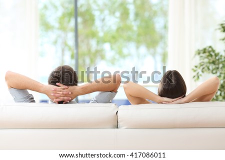 Rear view of a couple relaxing on a sofa at home and looking outside a green background through the window of the living room